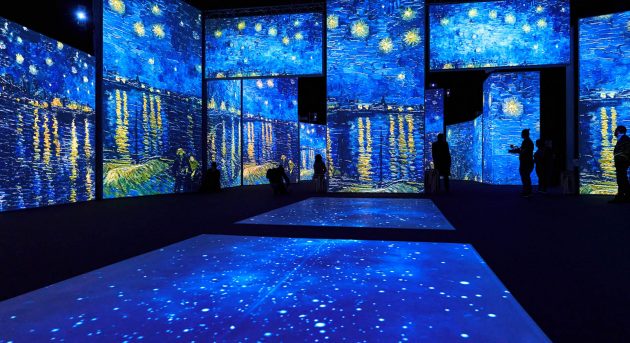 A dark room with multiple projections of Starry Nights by Vincent Van Gogh on the walls and floor. There are blue night skies with swirly stars