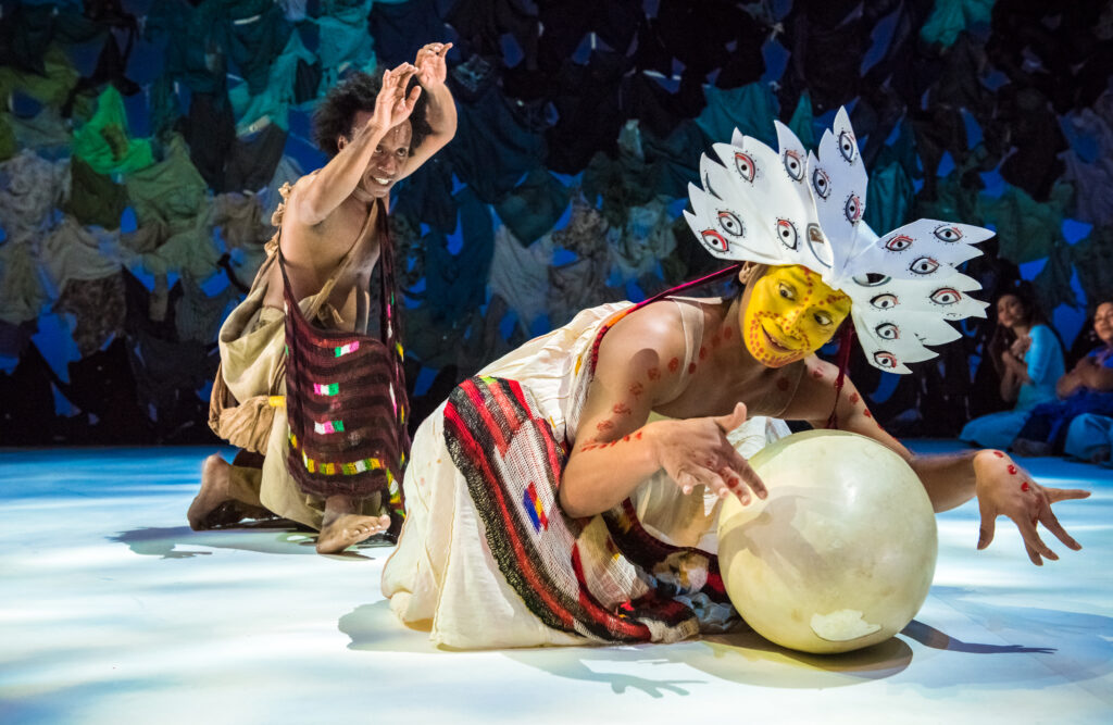 Indonesian Dancer Rianto performs in a stylised costume representing a bird with bright yellow face paint and protects a large egg. In the background the "The man who saves the world" can be seen with a bag slung across his chest and arms above head.